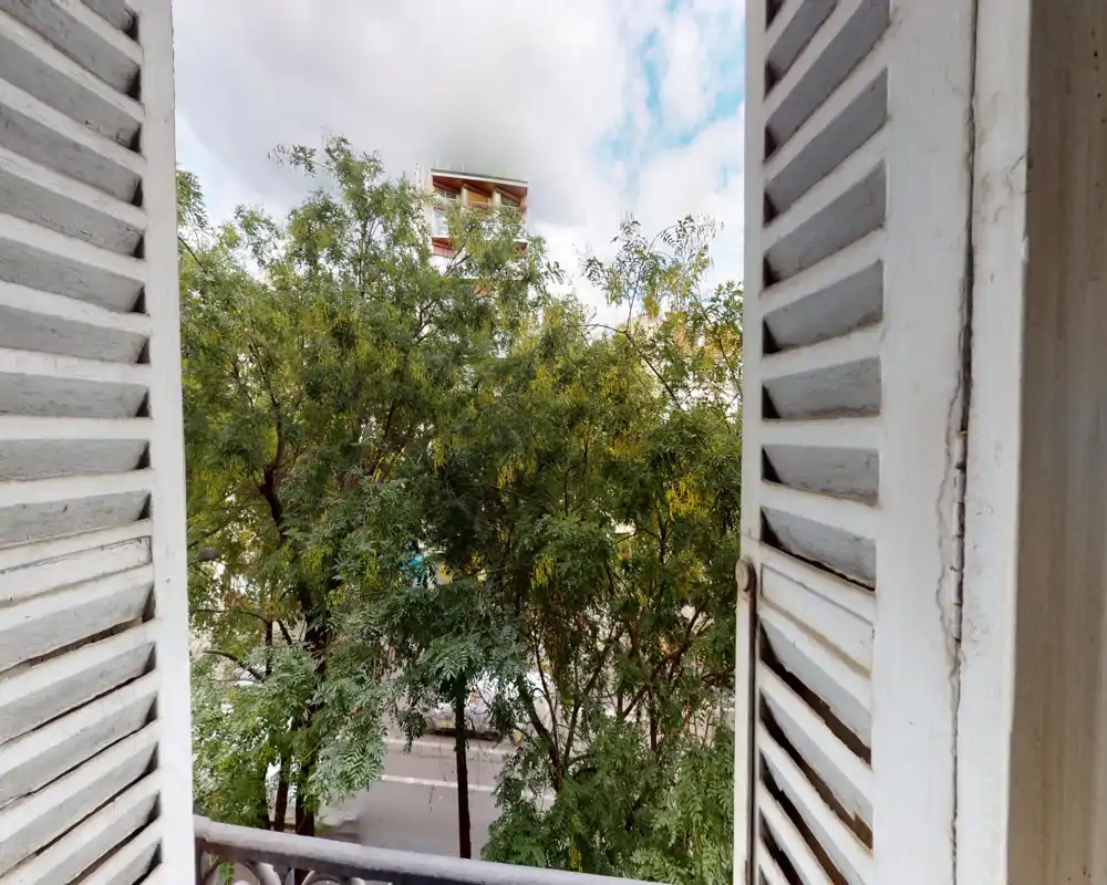 Vue depuis un petit balcon encadré de volets blancs, donnant sur des arbres feuillus et une rue calme — panorama urbain lumineux et apaisant.