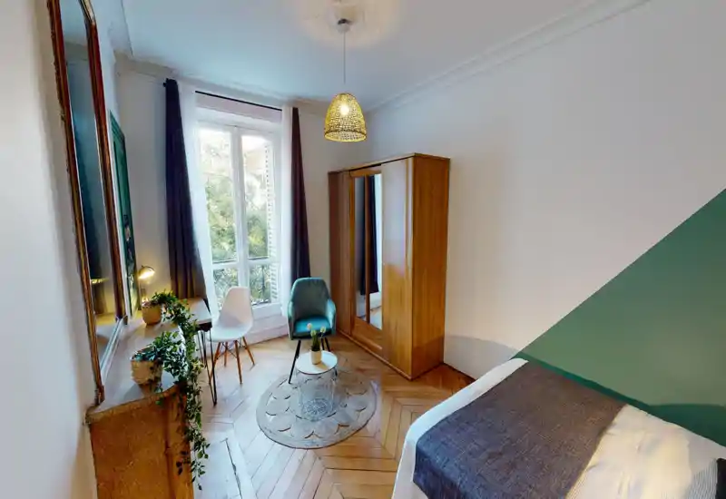 Chambre lumineuse et élégante avec lit visible, parquet, armoire avec miroir, fauteuil confortable et lumière naturelle provenant d'une grande fenêtre.