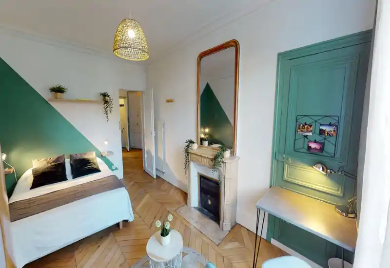 Chambre lumineuse et élégante avec grand lit double contre un mur vert, parquet en chevrons, cheminée décorative et petit bureau près de la fenêtre — chaleureuse et bien éclairée.