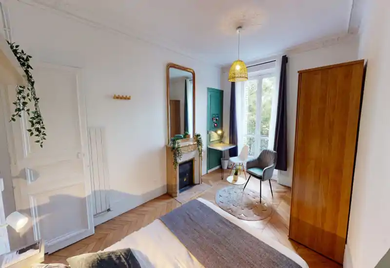 Chambre lumineuse et soignée avec un lit au premier plan, parquet, cheminée décorative et une grande fenêtre apportant beaucoup de lumière naturelle.