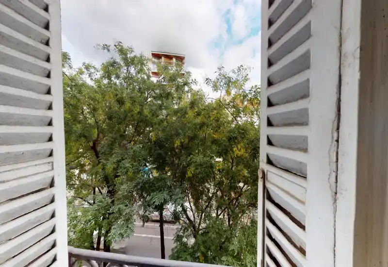 Vue depuis un petit balcon encadré de volets blancs, donnant sur des arbres feuillus et une rue calme — panorama urbain lumineux et apaisant.