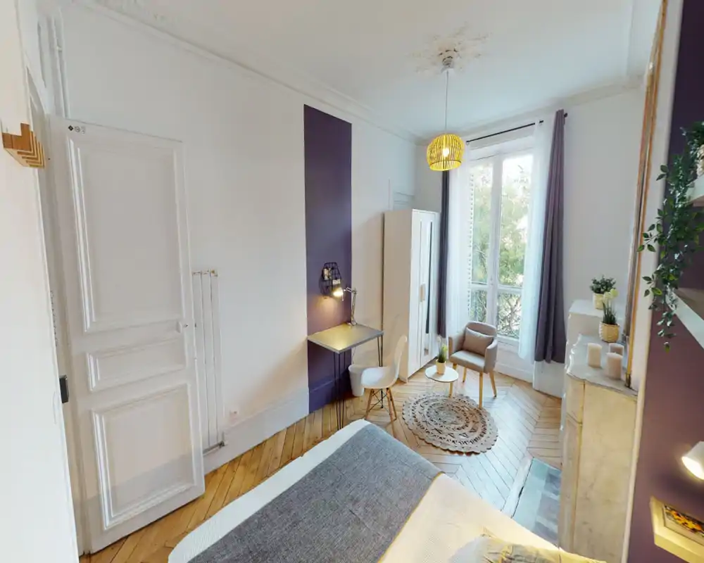 Bright, stylish bedroom with a large window, decorative rug and a comfortable bed in the foreground. The room features a small desk, wardrobe and seating area, offering a cozy, well-lit space ideal for relaxing or working.