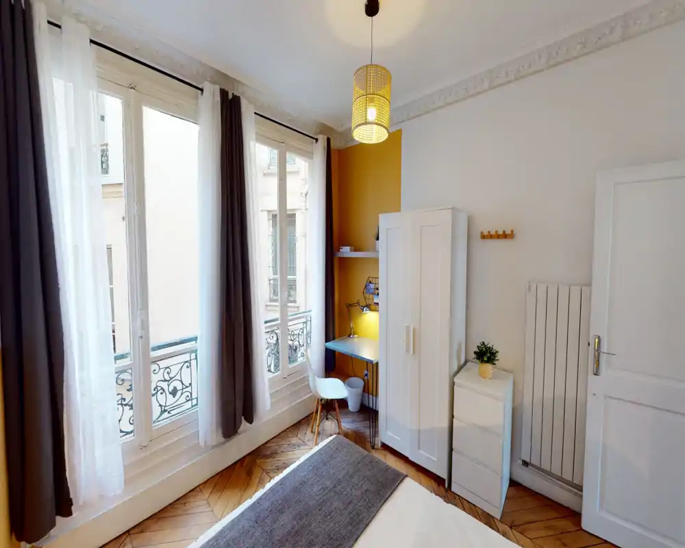 Chambre lumineuse et élégante avec grandes fenêtres, lit visible au premier plan, coin bureau compact et armoire — idéale pour une personne ou un couple.