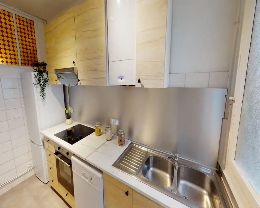 Cuisine moderne et compacte avec meubles en bois clair, plan de travail effet marbre, évier double en acier inoxydable et plaque à induction. Des bocaux décoratifs et une petite plante apportent une touche chaleureuse.