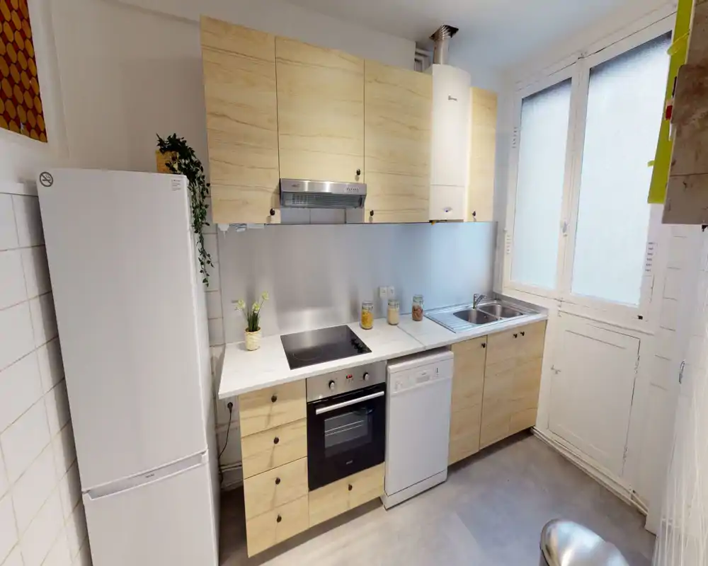 Cuisine compacte et moderne avec placards en bois clair, plaque à induction, four encastré, lave-vaisselle et évier double près d'une grande fenêtre dépoli — lumineuse et bien rangée pour cuisiner.