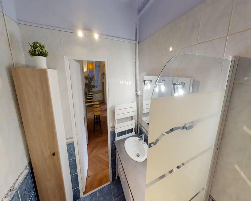 Salle de bain compacte et bien éclairée avec lavabo, miroir, radiateur porte-serviettes et pare-douche en verre dépoli ; murs carrelés et armoire en bois.
