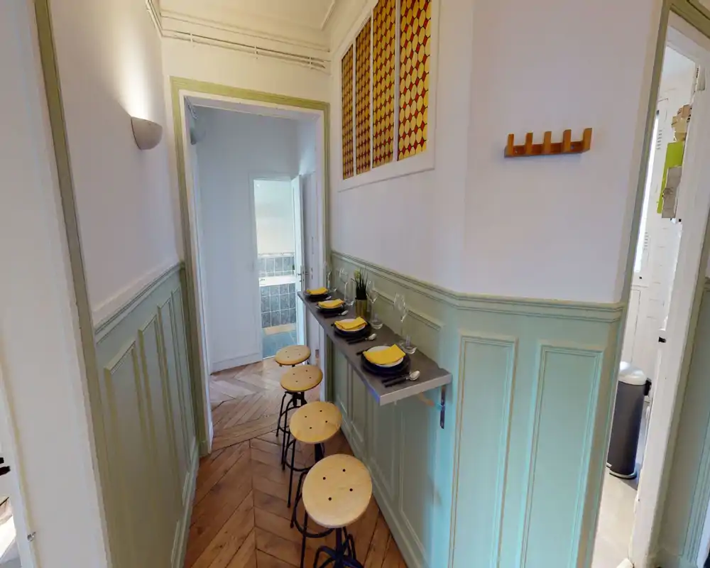 Coin repas compact dans le couloir avec un plan de travail mural et quatre tabourets en bois, dressé avec assiettes et verres — parfait pour un repas rapide ou le petit-déjeuner.