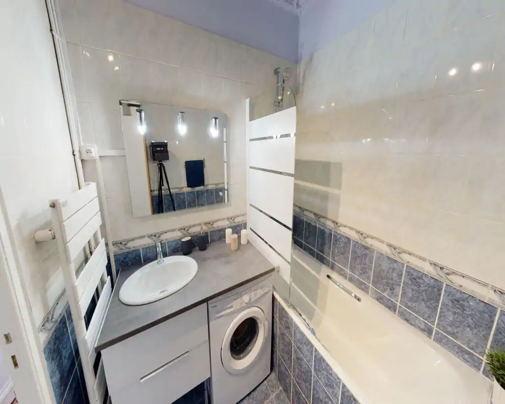 Salle de bain soignée avec vasque intégrée au plan, baignoire avec paroi vitrée et machine à laver sous le plan. Carrelage blanc et bleu pour une apparence propre et fonctionnelle.