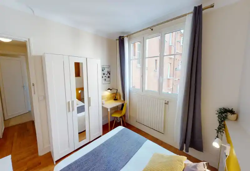 Chambre lumineuse et bien décorée avec grande fenêtre, lit confortable avec coussins et plaid, armoire et petit bureau près de la fenêtre — idéale pour un séjour citadin cosy.