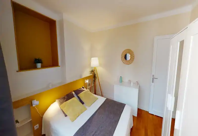 Chambre lumineuse et soignée avec grand lit double, tête de lit jaune, lampe d'appoint et décorations élégantes pour une ambiance chaleureuse.