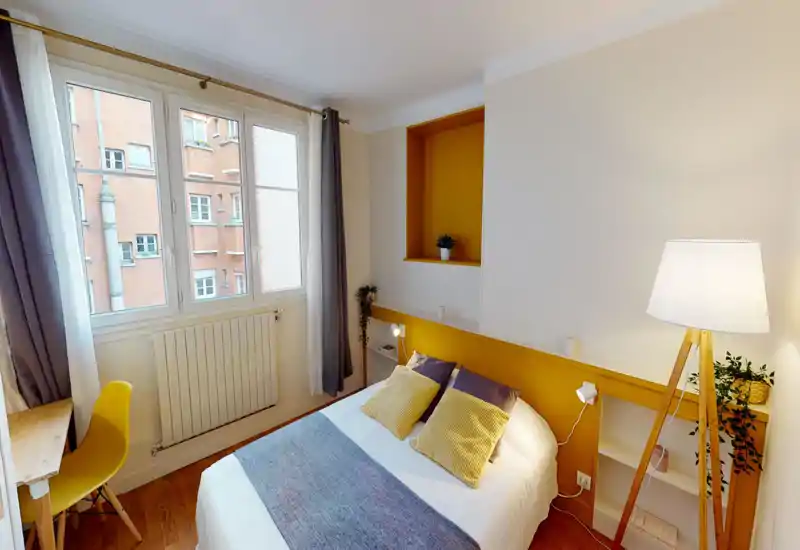 Chambre double lumineuse et bien décorée avec un mur jaune chaleureux, coussins et textiles confortables. Grande fenêtre apportant la lumière naturelle et un petit espace de travail près de la fenêtre.