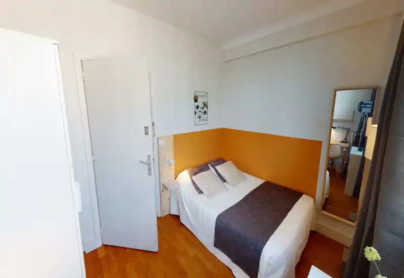 Chambre lumineuse et soignée avec un lit double contre un mur accent orange, lampe de chevet et décorations simples. Sol en bois et miroir en pied reflétant un petit espace de travail.
