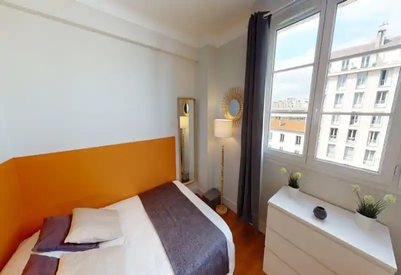 Chambre lumineuse et chaleureuse avec lit double, coussins décoratifs, lampe sur pied et commode près d'une grande fenêtre offrant une vue sur la ville.