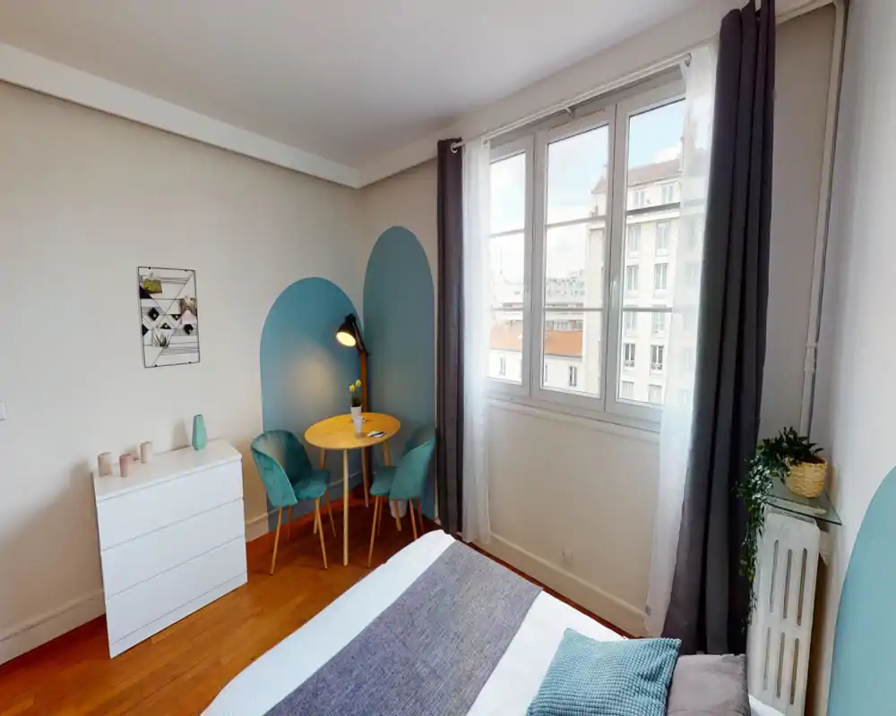 Bright, styled bedroom with a visible double bed in the foreground, a small round table with two teal chairs by the window, and soft decorative accents.