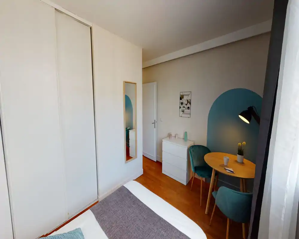 Bright, well-styled bedroom with a visible double bed in the foreground, a white chest of drawers, teal accent chairs and a small round dining table by the window.