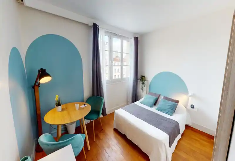 Bright, well-styled bedroom with a double bed, teal accent headboard, wooden floor and a small dining/work table by the window.