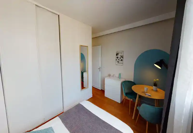 Bright, well-styled bedroom with a visible double bed in the foreground, a white chest of drawers, teal accent chairs and a small round dining table by the window.