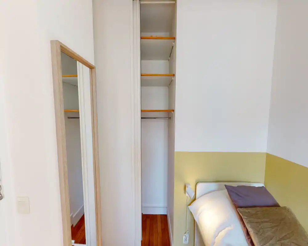 Camera da letto piccola e luminosa con letto singolo parzialmente visibile, lampada da comodino e armadio a muro aperto con ripiani. Pareti bianche con accento giallo caldo e pavimento in legno per un'atmosfera accogliente e minimale.