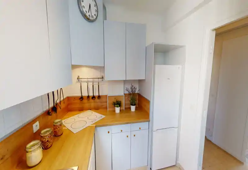 Bright, compact kitchen with wooden countertops, white cabinets and a fridge; utensils hang on a rail and decorative jars and plants add a cozy touch.