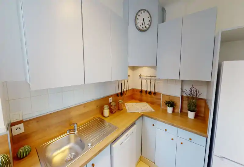 Compact modern kitchen with white cabinets, warm wooden countertops, built-in sink and induction hob — bright and neatly organized, with small plants and jars adding a cozy touch.