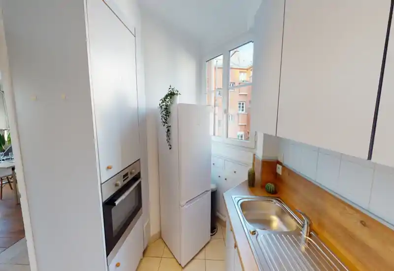 Compact, bright kitchen with white cabinets, a freestanding refrigerator, built-in oven and a wooden countertop with stainless sink; natural light from the window and small plants create a pleasant, lived-in feel.