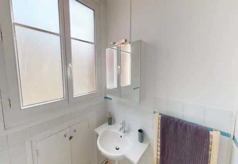 Small bright bathroom with a wall-mounted sink, mirrored cabinet and frosted window allowing natural light. Clean white tiles and a towel radiator create a neat, functional space ideal for a rental listing.