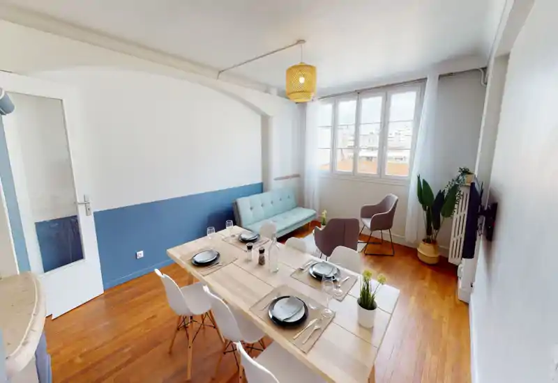 Bright, well-styled living/dining area with a wooden dining table set for four, a light-blue sofa and comfortable armchairs near a large window letting in natural light.