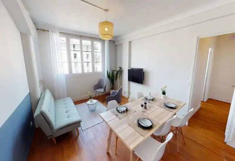 Bright, styled living room with a small dining table set for four. The space features a light sofa, two accent chairs, a wall-mounted TV and large windows that flood the room with natural light — ideal for socializing or remote work.
