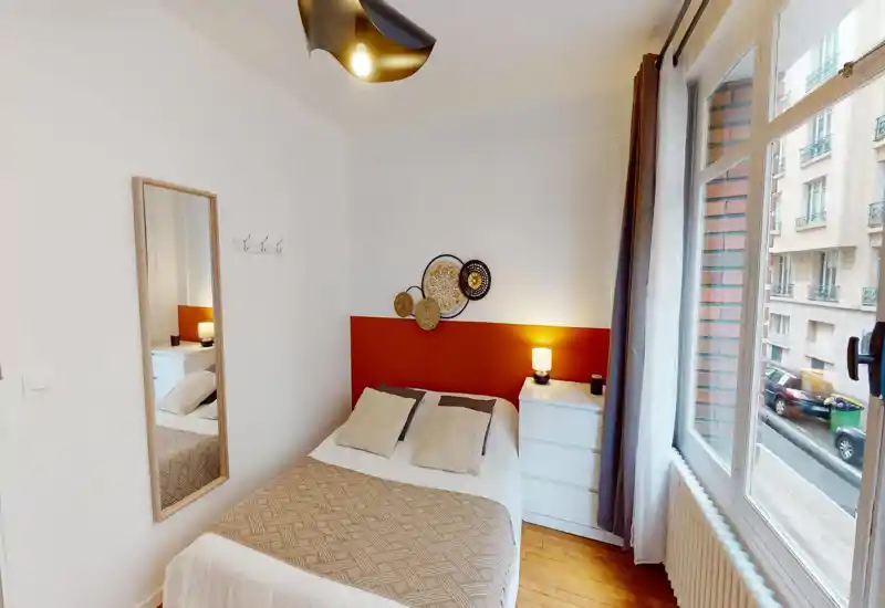 Chambre claire et chaleureuse avec un lit double bien fait, tête de lit décorative et lampe de chevet. Grande fenêtre apportant une belle lumière naturelle et parquet chaleureux — idéale pour une annonce locative.