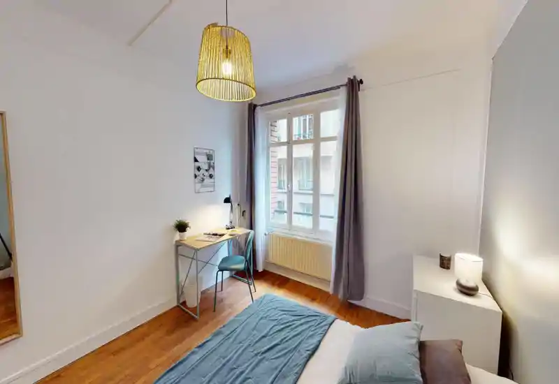 Bright, neatly staged bedroom with a made bed, bedside table and a small desk by the window; warm pendant lighting and wooden floors create a cozy atmosphere.
