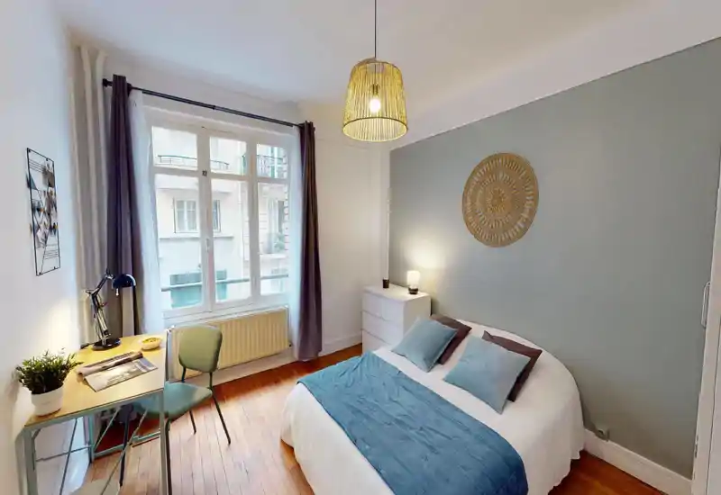 Well-lit bedroom with a double bed dressed in blue accents, bedside chest and a small desk by the window — cozy, modern styling and large window provide pleasant natural light.