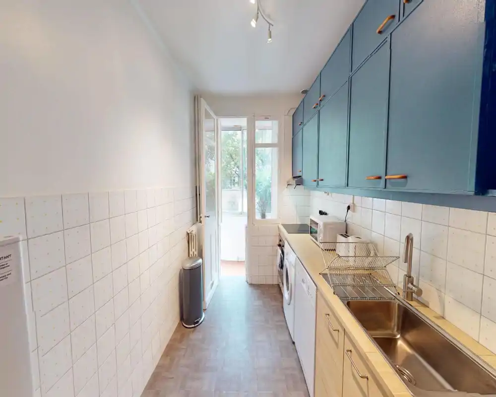 Cuisine en longueur lumineuse avec placards supérieurs bleus, plans de travail en bois, évier en inox et appareils électroménagers ; une porte/fenêtre à l'arrière apporte une belle lumière naturelle.