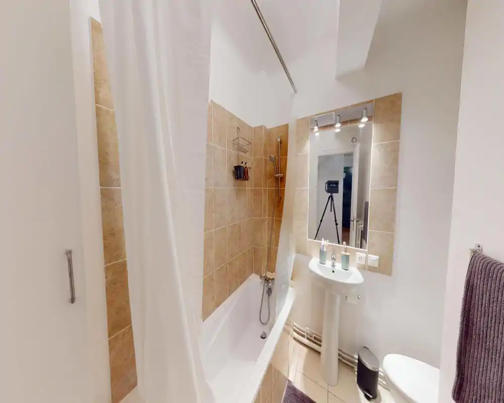 Salle de bains propre et lumineuse avec baignoire/douche, lavabo sur pied, miroir éclairé et équipements modernes.