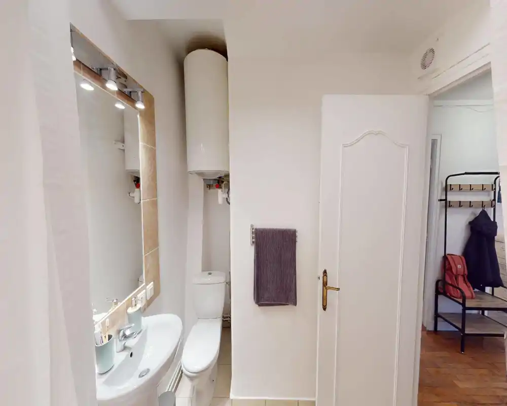 Salle de bain compacte et bien éclairée avec lavabo, miroir à éclairage intégré, WC et chauffe-eau mural. Carrelage neutre et porte-serviette rendent l'espace fonctionnel.