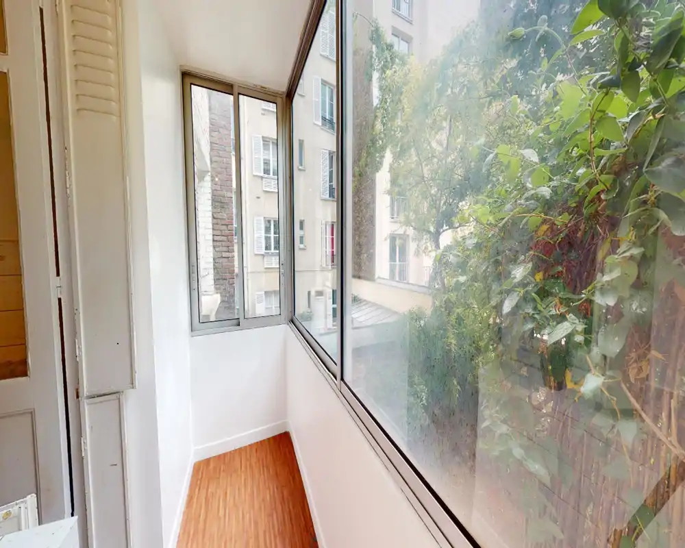 Balcon fermé / véranda avec grandes fenêtres donnant sur une cour végétale. Sol en bois étroit, murs blancs et bonne luminosité naturelle pour cet espace de transition.