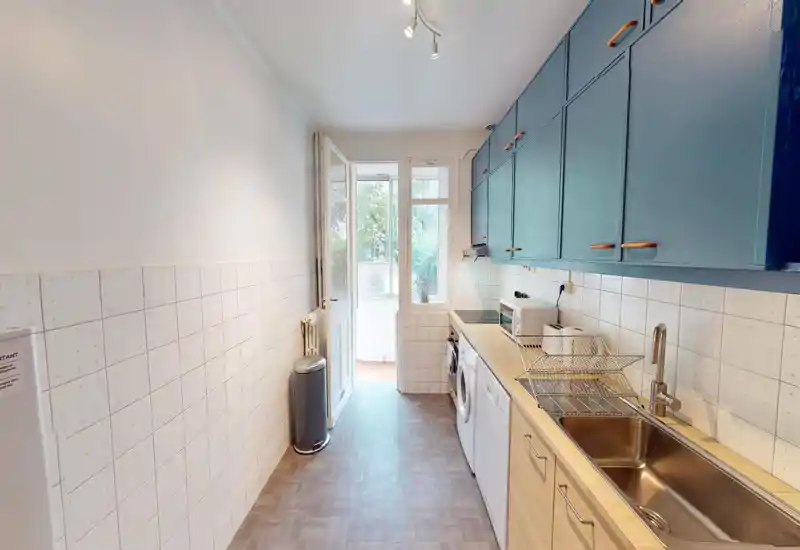 Bright galley kitchen with blue upper cabinets, wooden countertops, stainless sink and appliances; natural light from a door/window at the far end creates an airy feel.