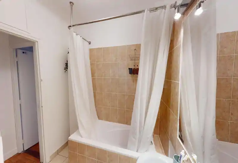 Clean tiled bathroom with a bathtub and shower curtain, wall-mounted toiletries and a large mirror visible — bright, functional space.
