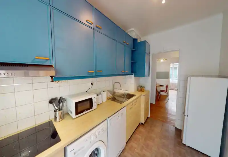 Bright, compact kitchen with blue upper cabinets, a wooden countertop, built-in sink and appliances including stove, microwave, dishwasher and washing machine. Practical layout with a clear work surface and pleasant color contrast.