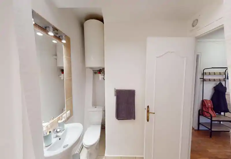 Compact, well-lit bathroom with a sink, mirror with integrated lights, toilet and a wall-mounted water heater. Clean neutral tiles and a towel rack make it functional for guest use.