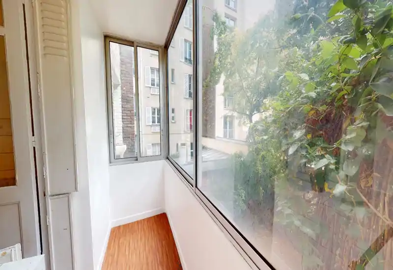 Enclosed balcony / sunroom with large windows looking onto a leafy courtyard. Narrow wooden floor, white-painted walls and plenty of natural light make it a bright transitional space.