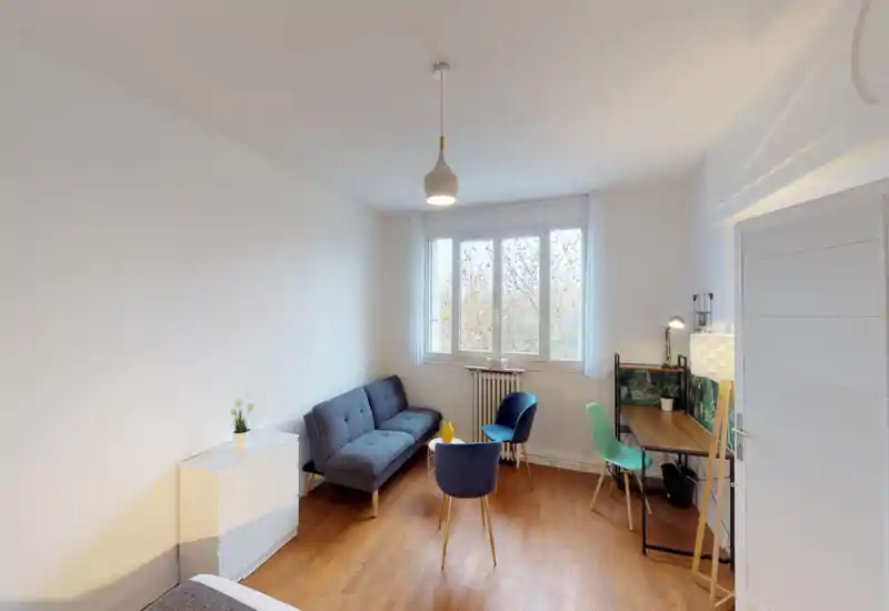 Angolo camera luminoso e minimal con piccolo divano, letto parzialmente visibile in primo piano, finestra che offre luce naturale e una piccola scrivania — ideale per un monolocale o un singolo in affitto.