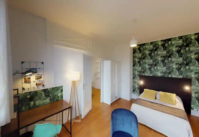 Camera da letto luminosa e di stile con letto matrimoniale davanti a una parete decorativa verde a foglie, lampade ai lati e cuscini gialli accoglienti. Pavimento in legno, armadio bianco e una scrivania rendono lo spazio pratico e ben arredato.