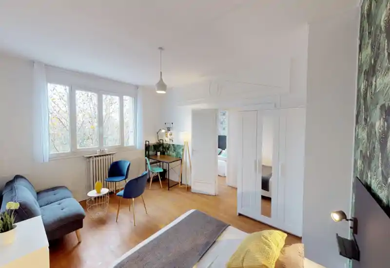 Camera da letto luminosa e ben allestita con letto comodo in primo piano, area salotto accogliente e una piccola scrivania vicino alla finestra. Le ampie finestre e il pavimento in legno creano un ambiente arioso e invitante, perfetto per annunci di affitto.