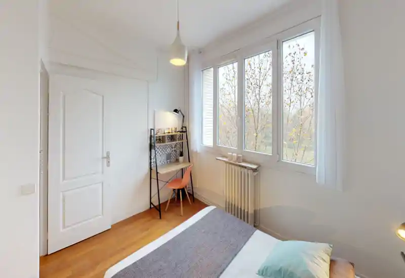 Chambre lumineuse et minimaliste avec lit bien fait, grandes fenêtres apportant une belle lumière naturelle et un petit coin bureau — idéale pour se reposer et travailler.
