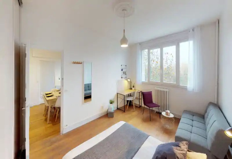 Chambre lumineuse et moderne avec lit au premier plan, petit bureau près de la fenêtre et canapé compact — parquet chaleureux et larges fenêtres apportant une belle lumière naturelle.