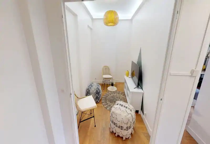 Petit espace salon aménagé avec chaises en rotin, poufs texturés, tapis à motifs et meuble TV ; murs blancs et sol en bois chaleureux pour une atmosphère moderne et cosy.