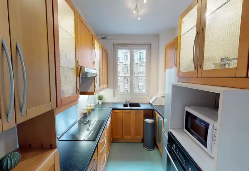 Cuisine compacte et lumineuse avec des placards en bois chaleureux, plans de travail noirs, électroménagers encastrés et une fenêtre apportant de la lumière naturelle — espace propre et fonctionnel adapté aux appartements urbains.