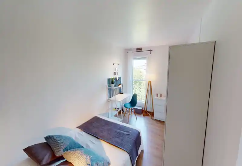 Chambre lumineuse et bien mise en scène avec un lit au premier plan, un petit bureau près de la fenêtre et une grande armoire. Textiles doux et lampadaire apportent une ambiance chaleureuse, idéale pour une annonce locative.