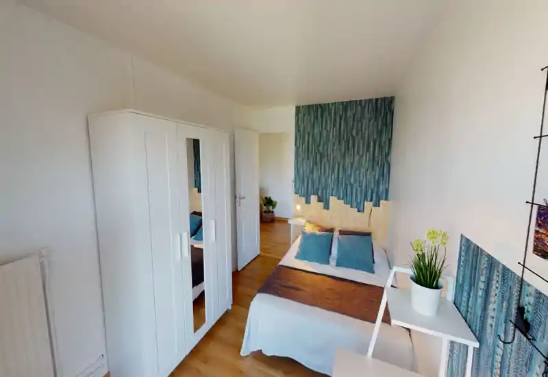 Chambre lumineuse et bien décorée avec un lit double, une tête de lit à motif et coussins assortis, une armoire blanche avec miroir et un petit bureau avec plante — idéale pour un séjour confortable.