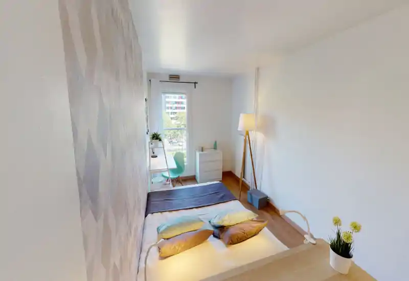 Chambre lumineuse et soignée avec un lit bas garni de coussins et d'un plaid, un petit bureau près de la fenêtre et une lampe sur pied apportant une lumière chaleureuse — idéale pour une location cosy et compacte.